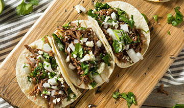 Three tacos filled with meat, cilantro, onions, and cheese on a wooden board.